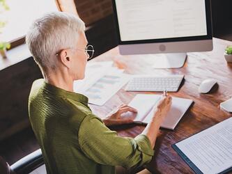 mature businesswoman writes in notepad at wooden desk in front of computer, surrounded by documents